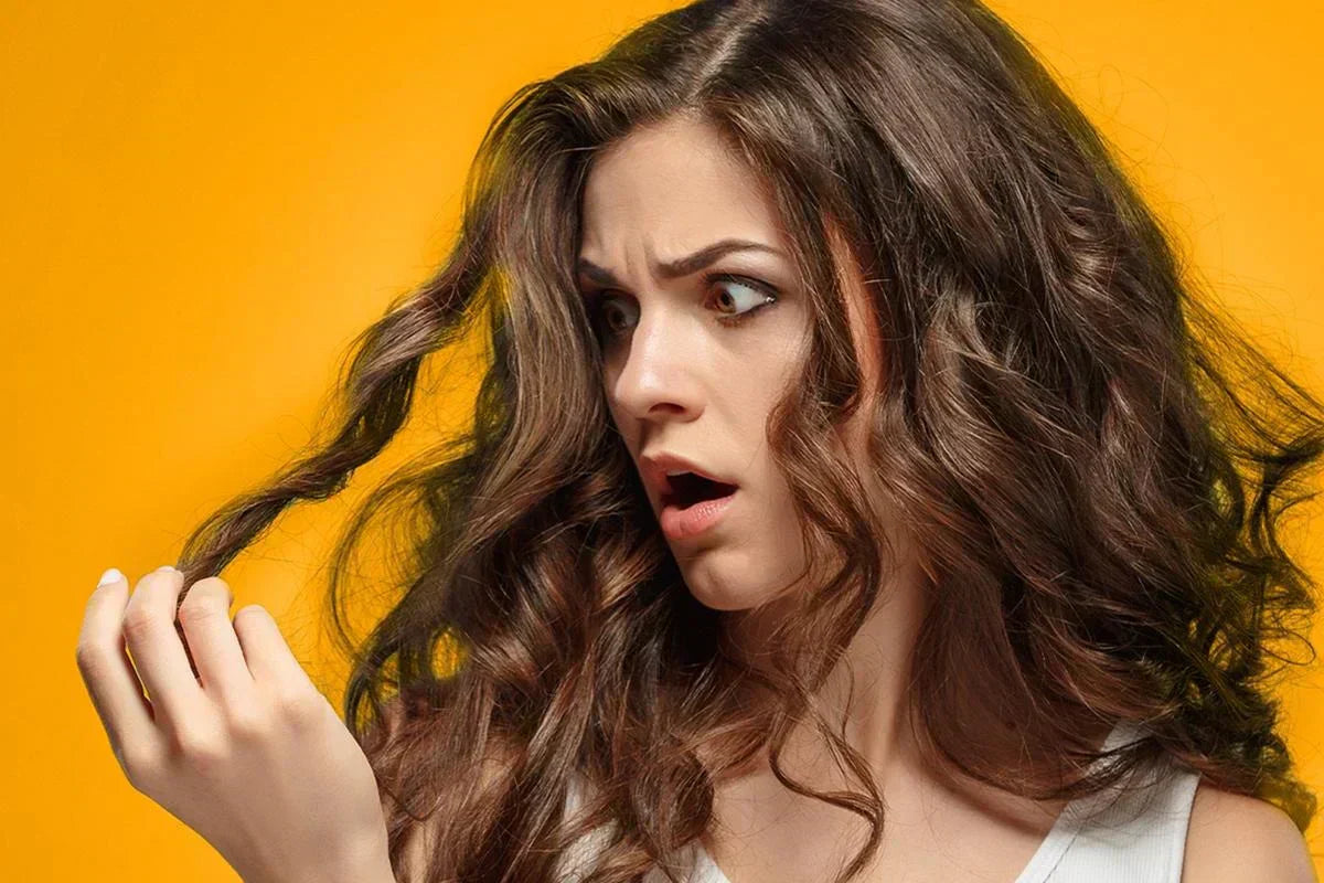 A person with long, wavy brown hair playfully curls a strand of hair with their fingers against a bright yellow background.