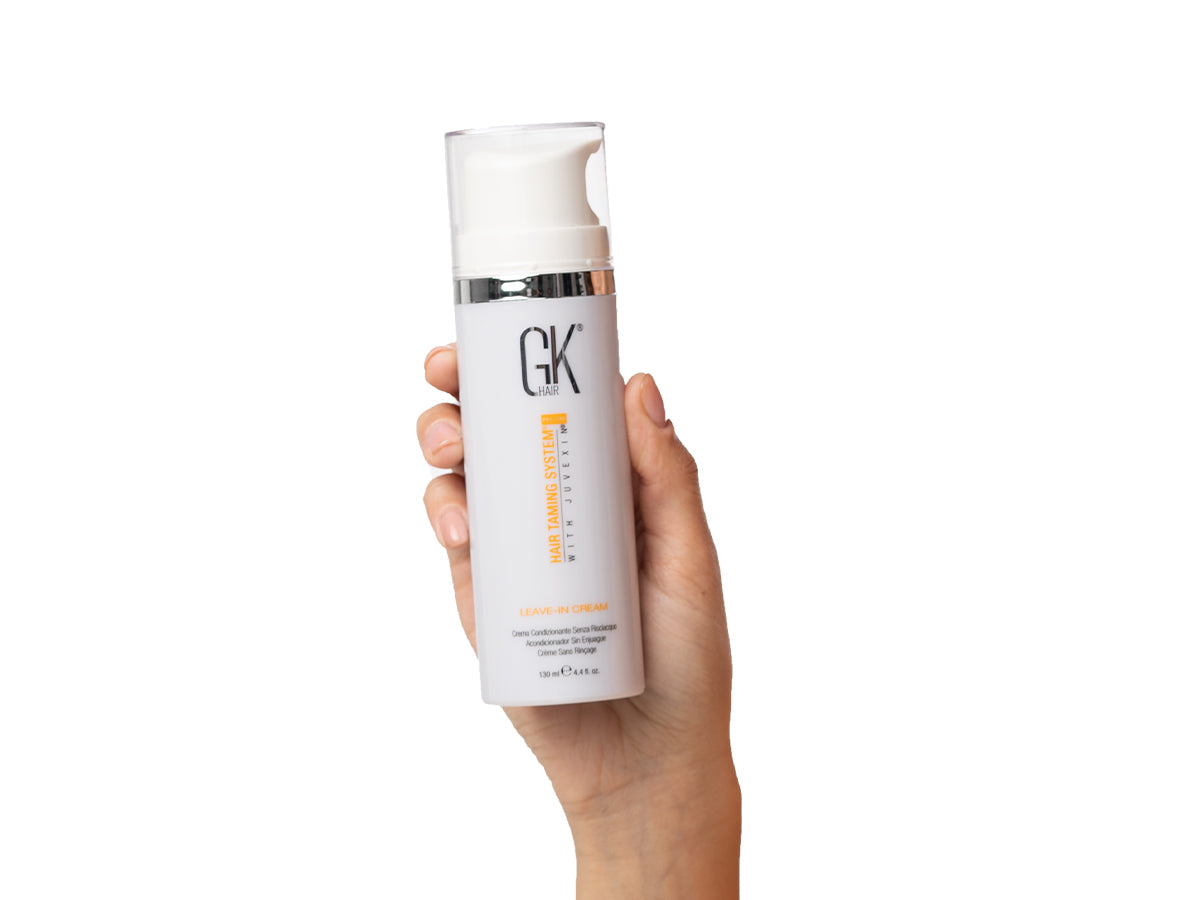 A hand holds a bottle of GK Hair leave-in cream, featuring a sleek design with a pump top and branding prominently displayed.