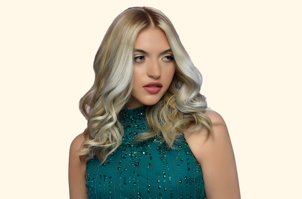 A woman with long, wavy blonde hair wears a sparkling emerald green dress with intricate beadwork, set against a light beige background.